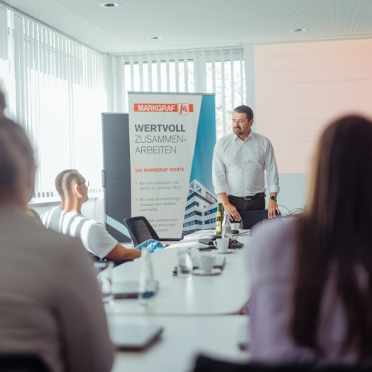 Ein Mann im weißen Hemd hält in einem modernen Konferenzraum einen Vortrag vor sitzenden Mitarbeitern, im Hintergrund ein Roll-up mit dem Markgraf-Slogan "WERTVOLL ZUSAMMENARBEITEN".