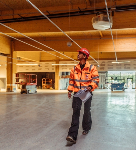 Polier oder Bauleiter der Firma Markgraf in oranger Warnjacke und rotem Schutzhelm hält Pläne in der Hand und geht durch einen noch im Ausbau befindlichen, großen Innenraum mit Holzdachkonstruktion.