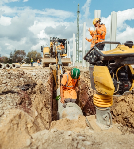 Bauarbeiter verlegen in einem tiefen Graben ein großes Betonrohr. Ein Kollege telefoniert, und eine gelbe Rüttelplatte steht im Vordergrund auf der Baustelle.