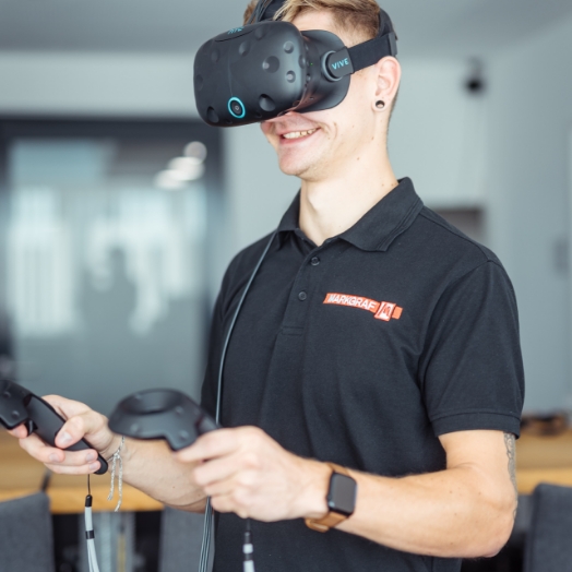 Lächelnder junger Mitarbeiter der Firma Markgraf mit schwarzem Poloshirt und Firmenlogo benutzt eine schwarze VIVE Virtual-Reality-Brille und hält zwei Controller in den Händen.